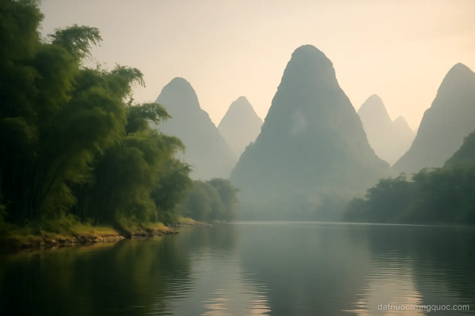 Karst limestone peaks rising beside a calm river with bamboo along the bank, hazy morning